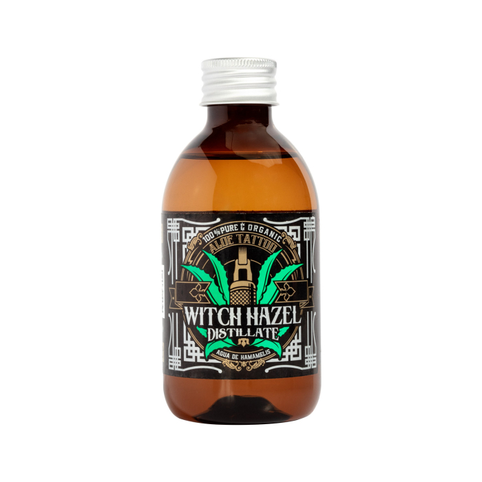 Un flacon en verre brun avec bouchon argenté, étiqueté « Aloe Tattoo Distillat d’hamamélis 250 ml », portant une étiquette noire avec détails verts et dorés, mentionnant « 100 % Pur & Bio » et « Aloe de Hamam ».